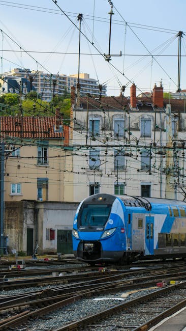 This image features overhead electrical wires and a modern blue and silver train moving along railway tracks in an urban setting. Behind the train, there are old and new residential buildings with red