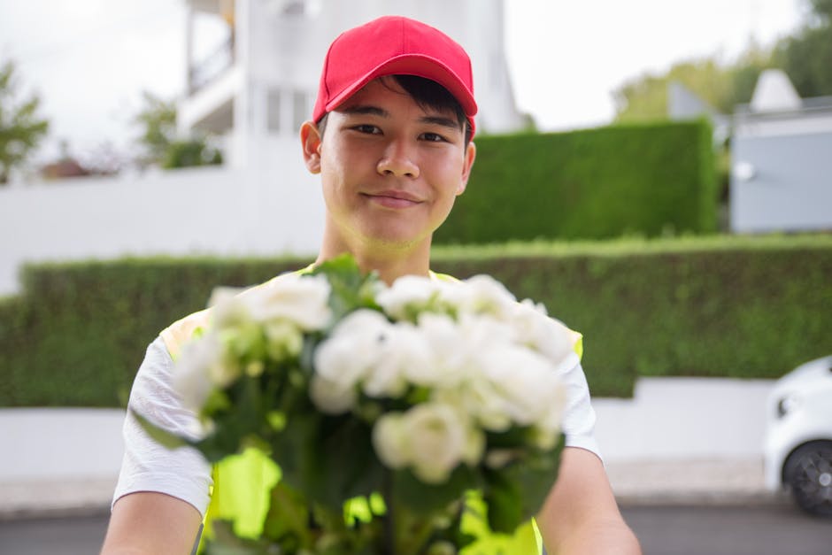 A delivery person wearing a red cap and a high-visibility yellow vest is holding out a bouquet of fresh, white flowers towards the camera. The bouquet includes roses and other mixed blooms wrapped in 